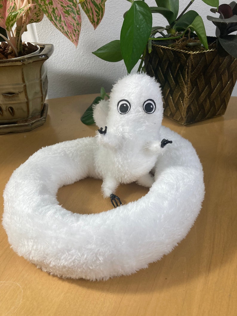 May include: A white plush toy shaped like a worm with a small, round body and a large, circular body. The toy has large, black eyes and small, black arms. The toy is sitting on a wooden surface, with plants in the background.