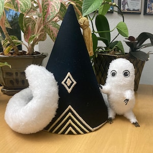 May include: A black, cone-shaped hat with gold trim and a tassel, next to a white plush donut and a small, white, furry creature with large eyes. The hat has a diamond and chevron design.