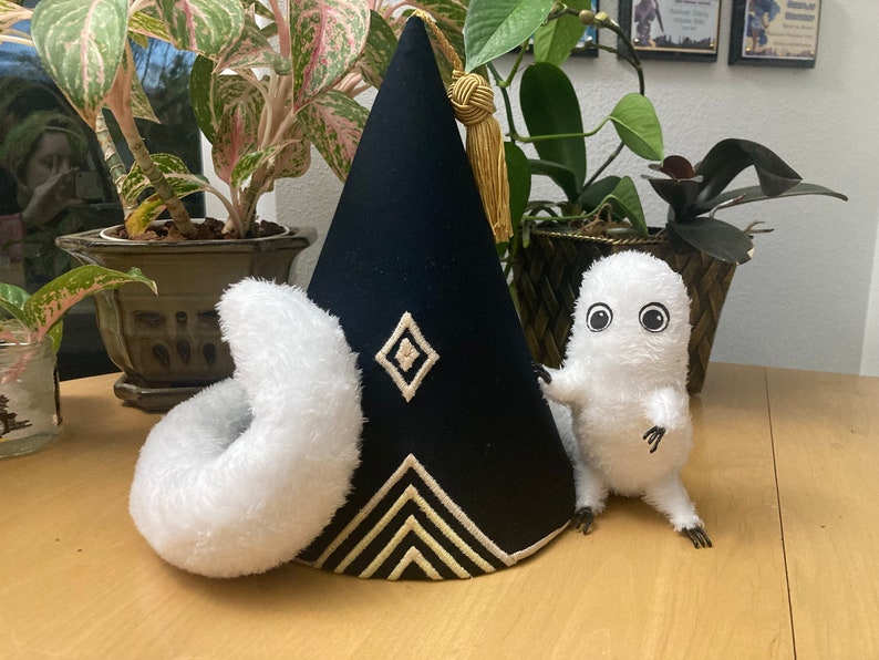 May include: A black, cone-shaped hat with gold trim and a tassel, next to a white plush donut and a small, white, furry creature with large eyes. The hat has a diamond and chevron design.