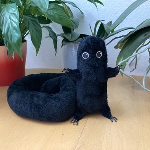 May include: A black plush creature with white-rimmed eyes and outstretched arms stands next to a matching black plush bed. The creature has small feet and a fluffy texture. The background includes potted plants on a light-colored wooden surface.