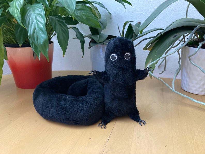 May include: A black plush creature with white-rimmed eyes and outstretched arms stands next to a matching black plush bed. The creature has small feet and a fluffy texture. The background includes potted plants on a light-colored wooden surface.