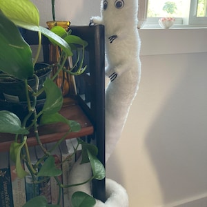 May include: A white plush creature with large eyes and small black claws clings to a dark wooden bookshelf. The creature has a long, cylindrical body and a curled tail. Green plants and books are visible.