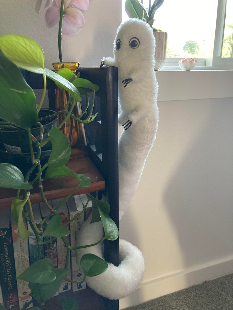 May include: A white plush creature with large eyes and small black claws clings to a dark wooden bookshelf. The creature has a long, cylindrical body and a curled tail. Green plants and books are visible.