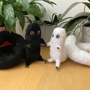 May include: Two plush toys, one black and one white, each with large, round eyes and small arms and legs. The black toy has a curled tail, while the white toy has a long, straight tail. The toys are set against a background of green plants.
