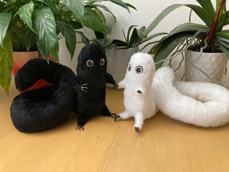 May include: Two plush toys, one black and one white, each with large, round eyes and small arms and legs. The black toy has a curled tail, while the white toy has a long, straight tail. The toys are set against a background of green plants.