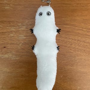 May include: A white plush keychain in the shape of a cartoonish creature with two black eyes and small black appendages. The keychain has a silver metal ring attached to the top.