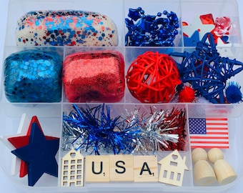 Puede incluir: Un contenedor de almacenamiento de plástico transparente lleno de suministros de artesanía de temática roja, blanca y azul para una actividad de temática patriótica. El contenedor incluye plastilina roja, blanca y azul, purpurina, estrellas y otras decoraciones.