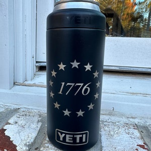 May include: A black Yeti Rambler 12 oz. can insulator with a silver lid. The insulator has a circle of stars around the number 1776. The Yeti logo is at the bottom of the insulator.