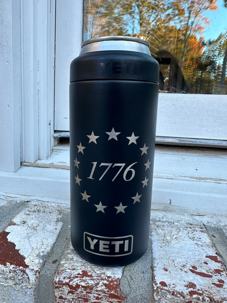 May include: A black Yeti Rambler 12 oz. can insulator with a silver lid. The insulator has a circle of stars around the number 1776. The Yeti logo is at the bottom of the insulator.
