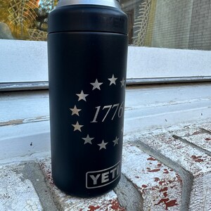 May include: Black Yeti Rambler with a silver inscription of a circle of stars and the number 1776. The Yeti logo is visible at the bottom of the can.