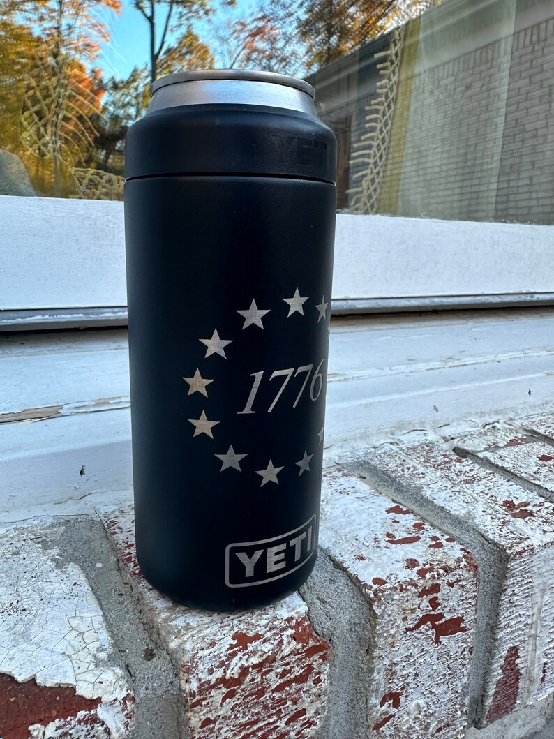 May include: Black Yeti Rambler with a silver inscription of a circle of stars and the number 1776. The Yeti logo is visible at the bottom of the can.