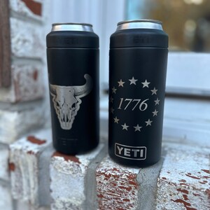 May include: Two black Yeti Rambler 12 oz. cans with different designs. The left can has a silver skull and horns design. The right can has a silver circle with 13 stars and the text "1776" inside. Both cans have the word "YETI" in silver lettering on the bottom.