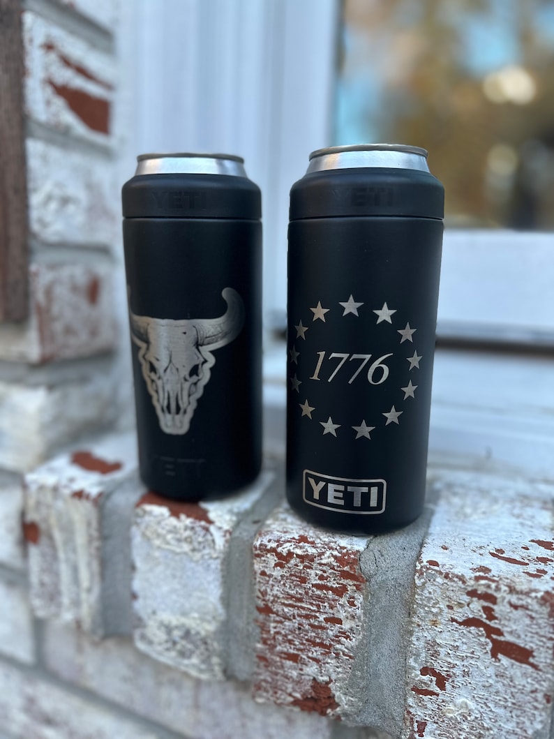 May include: Two black Yeti Rambler 12 oz. cans with different designs. The left can has a silver skull and horns design. The right can has a silver circle with 13 stars and the text "1776" inside. Both cans have the word "YETI" in silver lettering on the bottom.