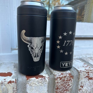 May include: Two black Yeti Rambler 12 oz. cans with engraved designs. The can on the left has a skull and horns design. The can on the right has a circle of stars with the number 1776 inside.