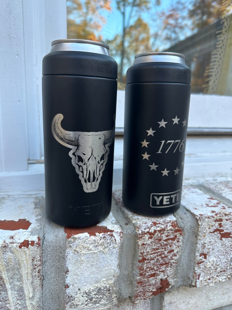 May include: Two black Yeti Rambler 12 oz. cans with engraved designs. The can on the left has a skull and horns design. The can on the right has a circle of stars with the number 1776 inside.