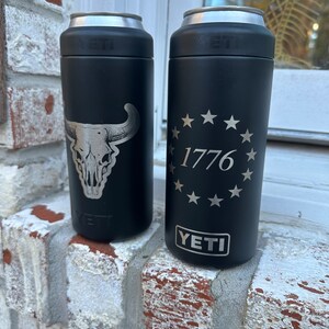 May include: Two black YETI Rambler 12 oz. cans with different designs. The can on the left has a silver skull and horns design. The can on the right has a silver circle with 13 stars and the text "1776" inside.