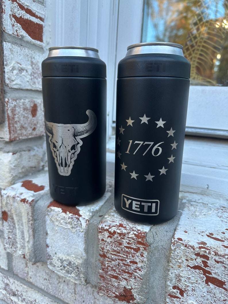 May include: Two black YETI Rambler 12 oz. cans with different designs. The can on the left has a silver skull and horns design. The can on the right has a silver circle with 13 stars and the text "1776" inside.