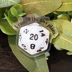 Peut inclure: Un dé à vingt faces blanc avec des chiffres noirs, lisant "J'ai ruiné les plans du mj", repose parmi des feuilles vertes et des fleurs blanches. Le dé est un accessoire de jeu, parfait pour les jeux de rôle.