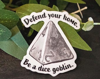 May include: A sticker featuring a grayscale D4 die with the text "Defend your home. Be a dice goblin." The sticker is surrounded by green leaves and white flowers. The workshop name "WANDERING BRUSH - WORKSHOP" is at the bottom.