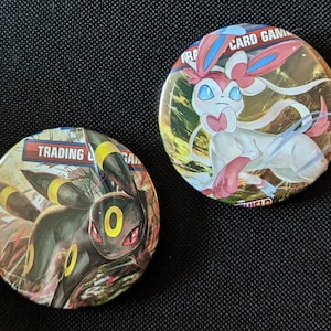 May include: Two round Pokemon Trading Card Game pins. One pin features a black and yellow Umbreon Pokemon. The other pin features a white and pink Sylveon Pokemon.