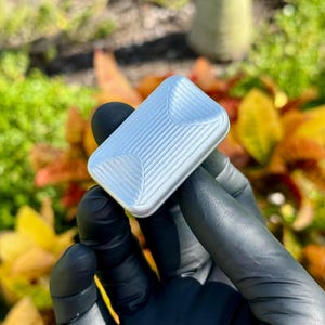 May include: A small, rectangular, silver-coloured metal object with a ribbed design. The object has rounded corners and is held in a gloved hand against a blurred background of green grass and colourful autumn leaves.
