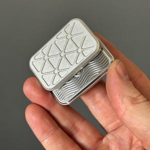 Op de afbeelding: Een zilverkleurig rechthoekig fidget-speeltje met een geometrisch ontwerp aan de bovenkant en een golvend patroon aan de binnenkant. De bovenkant heeft een driehoekig patroon met cirkels op de hoekpunten. Het fidget-speeltje wordt in een hand gehouden.
