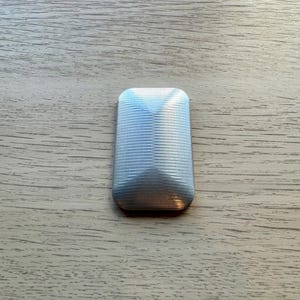 Może przedstawiać: Srebrna, prostokątna zabawka typu fidget o prążkowanej powierzchni i fazowanych krawędziach. Wierzch zabawki ma kształt piramidy. Zabawka spoczywa na jasnobrązowej drewnianej powierzchni.