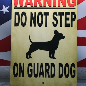May include: A yellow warning sign with black text that reads "WARNING DO NOT STEP ON GUARD DOG". A black silhouette of a small dog is in the center of the sign.