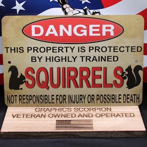 May include: A brown metal sign with the text "DANGER" in a red circle. The sign reads "This property is protected by highly trained squirrels. Not responsible for injury or possible death." The sign is on a wooden stand with the text "Graphics Scorpion Veteran Owned and Operated" and an American flag.
