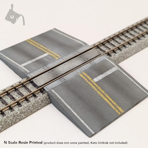May include: N scale model railroad crossing with gray asphalt road sections, yellow and white lane markings, and a metal crossing plate. The model railroad track runs through the center. The text "N Scale Resin Printed" is at the bottom.