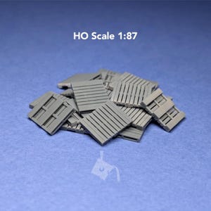 May include: A pile of small, gray, rectangular shipping pallets in HO scale 1:87. Each pallet features a slatted top surface and a structured base, designed for miniature modeling and dioramas, presented as a collection of individual units.