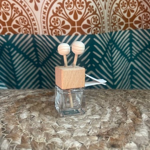 以下が含まれることがあります： 木製の蓋と2本の白いディフューザー棒が付いた、小さな透明なガラス瓶。瓶は織り目の表面の上に置かれています。