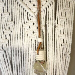 以下が含まれることがあります： 白いマクラメの壁掛けに、ドライフラワーが入った小さなガラス瓶と木製の栓が、茶色の革紐でぶら下がっている。