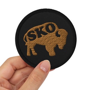 May include: A black round patch with a brown embroidered buffalo silhouette. The buffalo has the word "SKO" embroidered inside its outline.