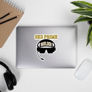 May include: Black and white sticker with a black silhouette of a person wearing a headset and sunglasses. The text "SKO PRIME" is above the silhouette in gold letters. The text "BOULDER" is on the headset in gold letters.