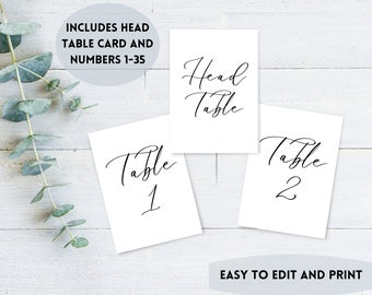 Op de afbeelding: Witte tafelkaarten met elegant zwart schrift. De kaarten bevatten "Head Table", "Table 1" en "Table 2". De afbeelding bevat ook de tekst "INCLUDES HEAD TABLE CARD AND NUMBERS 1-35" en "EASY TO EDIT AND PRINT".