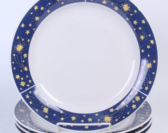 May include: Set of four white ceramic plates with a navy blue border. The border is decorated with gold stars and shooting star designs. The plates are stacked on a white metal plate holder.