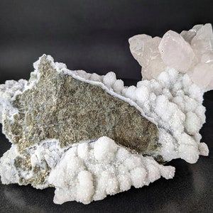May include: A cluster of white and pink quartz crystals on a gray rock. The crystals are clustered together in a variety of shapes and sizes. The rock is rough and textured.
