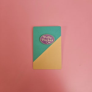 May include: Purple and gold enamel pin with the text "Polly Pocket" in a circle.