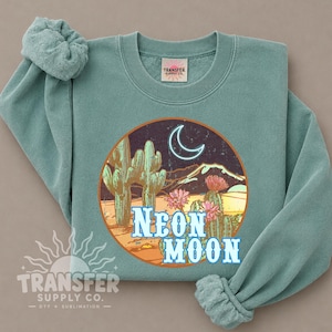 May include: A sage green crewneck sweatshirt featuring a circular graphic of a desert landscape at night with cacti, pink flowers, a glowing neon crescent moon, and the text NEON MOON in light blue. The garment has a ribbed collar and cuffs.