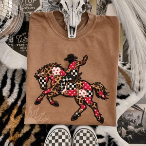 May include: A tan crewneck t-shirt featuring a central graphic of a person riding a horse. The design uses a patchwork style with leopard print, black and white checkerboard patterns, and red fabric with white hearts on both the horse and rider.