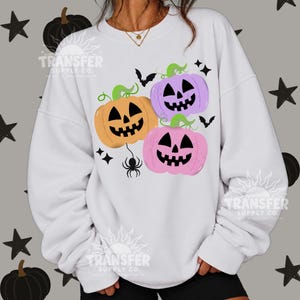 May include: A white crewneck sweatshirt featuring a graphic print of three jack-o'-lanterns in orange, purple, and pink. The design includes green stems, black bat silhouettes, a black spider hanging from a thread, and small black star shapes.