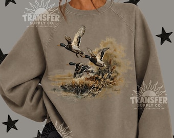 May include: A taupe crewneck sweatshirt featuring a graphic print of three mallard ducks in flight over a marshy landscape. The design includes detailed illustrations of the birds with brown, green, and white plumage, set against a soft, muted background.