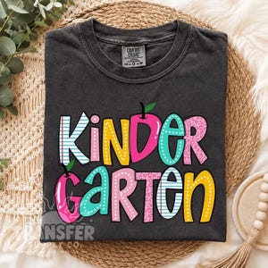 May include: A charcoal gray short-sleeve t-shirt featuring the word KINDERGARTEN in large, colorful, patterned letters. The design includes pink, yellow, teal, and blue colors with polka dot, leopard print, and lined paper patterns.