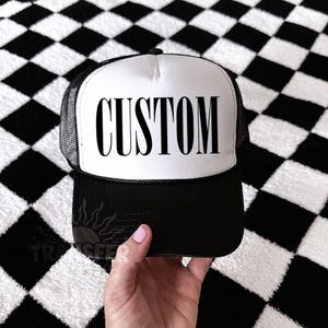 May include: A black and white trucker hat featuring a white front panel with the word CUSTOM printed in bold black capital letters, a black brim, and black mesh side and back panels. The hat has a structured front and a black plastic snapback closure.