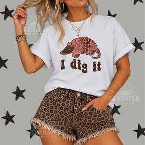 May include: A white short-sleeved t-shirt featuring a graphic of a pink and brown armadillo above the brown text "I dig it." The shirt is paired with brown and black leopard print denim shorts that have a frayed, raw-edge hemline.