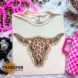 May include: Cream-colored t-shirt featuring a leopard print bull skull design with fringe and the text "TRANSFER SUPPLY CO." The shirt has a crew neck and short sleeves. The shirt is a Comfort Colors brand.