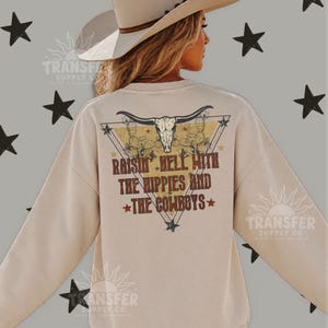 May include: A beige crewneck sweatshirt featuring a back graphic of a longhorn skull inside a triangle, surrounded by cactus illustrations and stars. The text reads, "RAISIN' HELL WITH THE HIPPIES AND THE COWBOYS" in a distressed, rust-colored font.