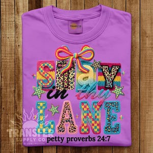 May include: A bright purple t-shirt featuring a colorful graphic with the text "Stay in thy LANE" and "petty proverbs 24:7". The design includes a multi-colored bow, stars, and letters with various patterns like leopard print and knit textures.