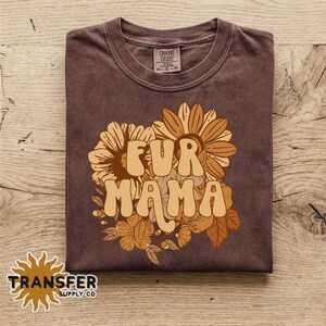 May include: A dark brown short-sleeve t-shirt featuring a graphic print of sunflowers and leaves in shades of yellow and orange. The text "FUR MAMA" is printed in a bold, rounded cream font across the center of the floral design.